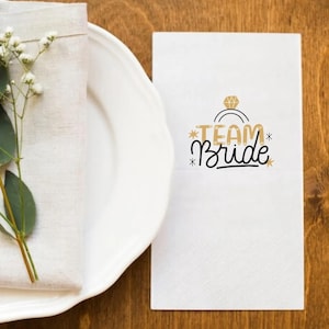 May include: White napkin with the text "Team Bride" in black lettering and a gold ring with a diamond graphic. The napkin is on a wooden table with a white plate and a white napkin with green flowers.