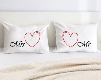 May include: Two white pillowcases with red hearts and the words "Mrs" and "Mr" printed on them. The pillowcases are on a white bed with a cream-colored blanket.