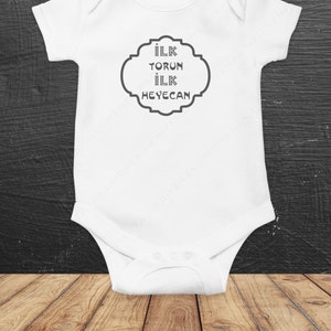 May include: White baby onesie with a gray design that says "ilk torun ilk heyecan" in a decorative frame.