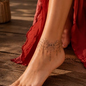 May include: A temporary tattoo applied to the ankle featuring a delicate black ink design of a central flower surrounded by intricate leafy vines and thin dangling chains with small geometric pendants that mimic the appearance of a fine jewellery anklet.