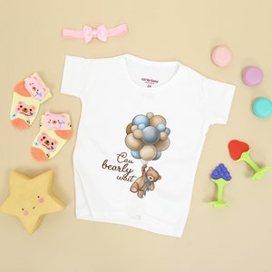 May include: White baby onesie with a brown bear holding balloons that say "Can bearly wait". The onesie is surrounded by a pink headband, yellow star, and colorful macarons.