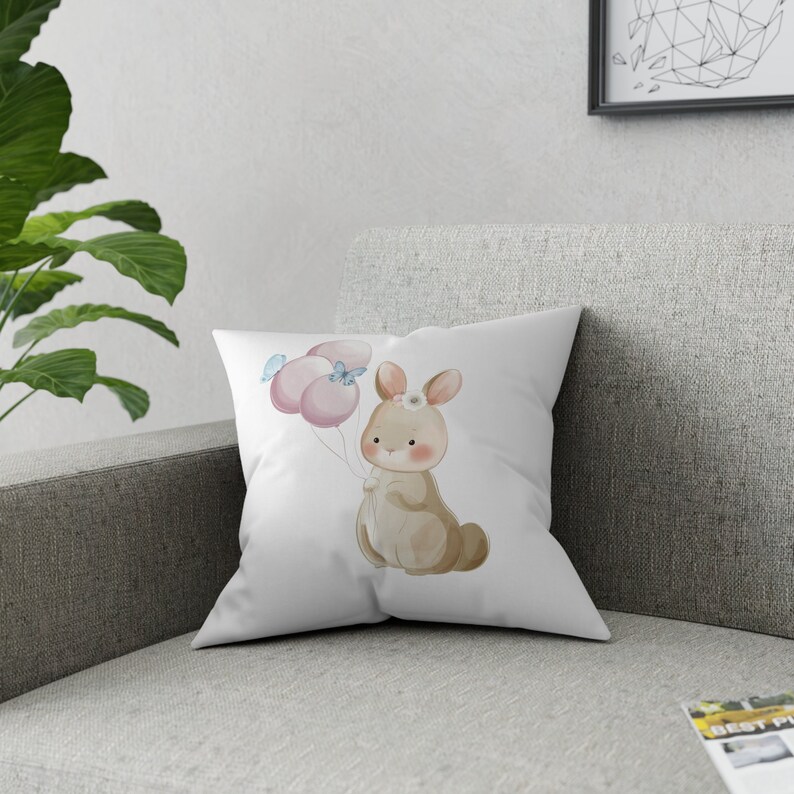 May include: A white throw pillow with a watercolor illustration of a brown bunny holding pink balloons with blue butterflies.