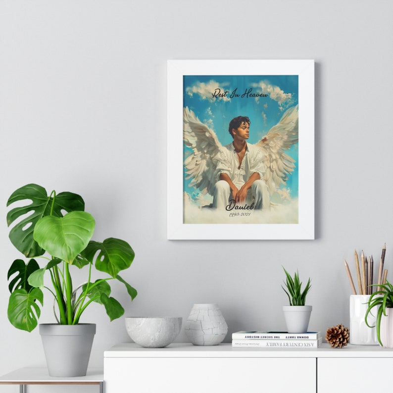 May include: A framed print with a white background and a blue sky with clouds. A man with brown skin and short black hair is sitting on a cloud with white angel wings. The text "Rest In Heaven" is at the top of the image and the text "Daniel 1998-2023" is at the bottom of the image.