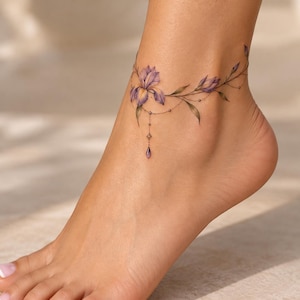 Puede incluir: Un delicado tatuaje de tobillo que presenta una flor de iris morada con hojas y tallos verdes, diseñado para parecerse a una fina tobillera. El tatuaje incluye líneas finas que cuelgan como cadenas con un pequeño dije morado en forma de gota.
