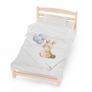 May include: A white and blue bunny with a pink flower on its head holds blue balloons with pink butterflies. The bunny is on a white blanket on a wooden bed frame.