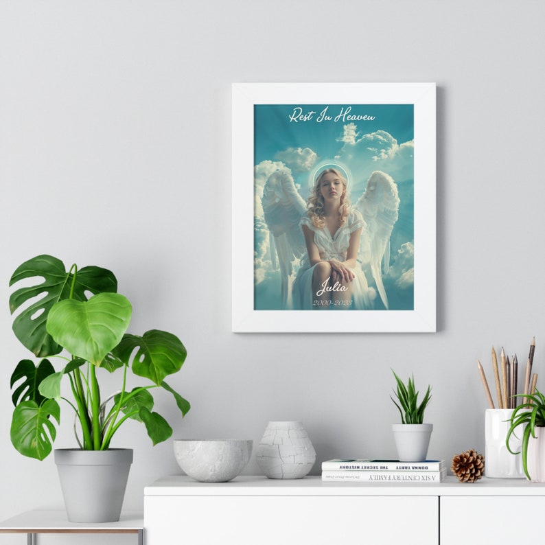 May include: A white framed print with a blue and white background featuring a young woman with angel wings sitting on a cloud. The text "Rest In Heaven" is at the top of the print and the text "Julia 2003-2023" is at the bottom.
