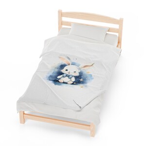 Pu&ograve; includere: Un letto bianco con una federa a motivi bianchi e blu con un coniglio dei cartoni animati con un papillon blu. La federa ha un effetto acquerello con stelle dorate.