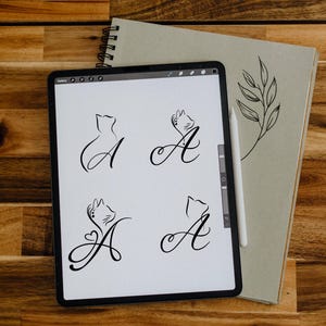 Pode incluir: Um tablet digital exibe quatro desenhos de arte de linha preta da letra "A" com ilustrações com tema de gato. Uma caneta e um caderno espiral com um desenho de folha estão ao lado do tablet em uma superfície de madeira.