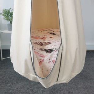 Puede incluir: Una silla colgante de lona blanca con ribete gris. La silla está colgada del techo y tiene un forro de tela estampado.