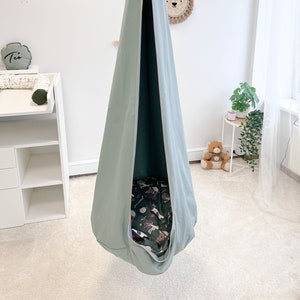 Puede incluir: Una silla colgante de color verde salvia con un ribete rosa claro. La silla está suspendida del techo y tiene un cojín estampado en su interior.