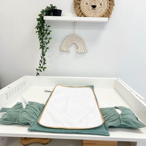 Può includere: Un fasciatoio bianco con un tappetino per il cambio verde, un asciugamano bianco e un porta fazzoletti in tessuto verde. C'è un giocattolo di legno sul fasciatoio e una scatola di legno sullo scaffale inferiore. Il fasciatoio è contro un muro bianco con uno scaffale sopra. Lo scaffale ha una testa di leone tessuta e un arcobaleno di macramé bianco appeso.