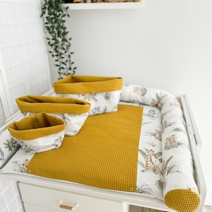 May include: A white changing table with a mustard yellow changing pad and three matching storage baskets. The changing pad has a white border with a jungle animal print. A decorative lion head and a toy are on the shelf above.