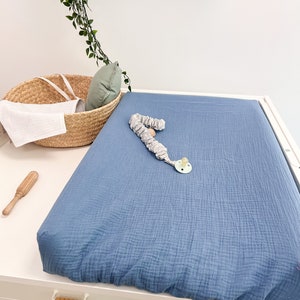 Peut inclure: Une housse de matelas à langer bleue sur une table à langer blanche avec un jouet en bois et un panier en osier avec une serviette blanche.
