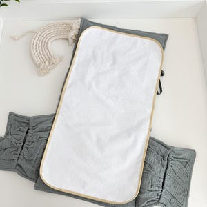 May include: A white baby changing pad with a tan border sits atop a gray changing pad cover. A decorative rainbow made of woven cream-colored material is in the upper left corner. The changing pad cover has a ruffled design.