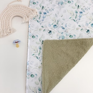 May include: A baby changing pad with a white floral print featuring blue and green leaves. The reverse side is a solid olive green. A beige macrame rainbow and a pacifier are also visible.