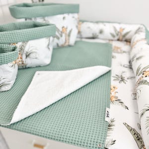 Peut inclure: Un ensemble d'articles en tissu gaufré vert sauge comprenant trois paniers de rangement, une housse de matelas à langer et un tissu plié. Les articles présentent un panneau en tissu blanc avec un motif imprimé de girafes et de feuilles tropicales. Une petite étiquette blanche indique Kals.