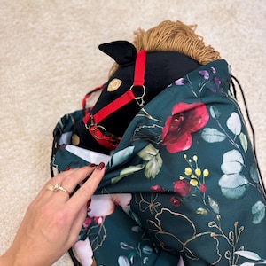 Könnte beinhalten: Ein schwarzes Steckenpferd mit rotem Zaumzeug schaut aus einem Kordelzugbeutel. Die Tasche ist dunkelgrün mit einem Blumenmuster aus roten Rosen, weißen Blüten und goldenen Akzenten. Das Pferd hat eine hellbraune Mähne.