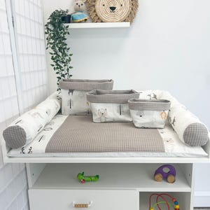 Pode incluir: Uma mesa de fraldas branca com um colchão de fraldas cinza e uma prateleira branca com uma cabeça de leão tecida pendurada na parede. O colchão de fraldas tem uma borda com padrão branco e cinza e uma textura de waffle cinza. Existem três cestas de armazenamento cinza com tecido estampado branco e cinza no colchão de fraldas. A mesa de fraldas tem três gavetas e uma prateleira inferior com um carro de brinquedo de madeira e um brinquedo de madeira com contas coloridas.