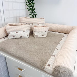 Op de afbeelding: Een aankleedstation voor baby's met een witte commode, een beige aankleedkussen en bijpassende accessoires. De accessoires omvatten vier stoffen opbergmanden met een wafeltextuur en een berenpatroon. De commode heeft drie lades.