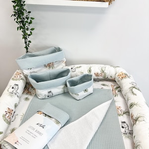 Puede incluir: Un juego de tres cestas de almacenamiento de tela verde salvia con textura de gofre y una manta de algodón doblada con una etiqueta que dice Kalis Cotton Blanket 100% organic cotton 70x100cm, dentro de un nido de bebé blanco con estampado.