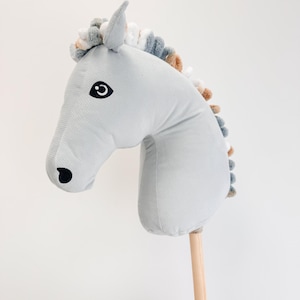 Puede incluir: Un caballo de balancín de peluche gris con un mango de palo de madera. El caballo tiene un ojo negro y una crin marrón y blanca.