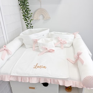 Peut inclure: Une table à langer blanche avec un matelas à langer, des coussins de protection et des paniers de rangement. Le matelas et les paniers sont ornés de nœuds roses. Le matelas à langer porte le nom "Lucia" brodé en fil doré. Une commode blanche avec des poignées en bois est en dessous.