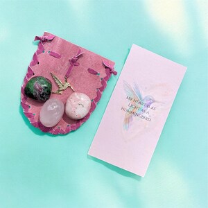 May include: A pink leather pouch with a gold hummingbird charm and three gemstones. A card with a hummingbird illustration and the text "MY HEART IS AS LIGHT AS A HUMMINGBIRD" is next to the pouch. The background is a light teal color.