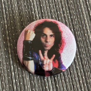 May include: A round button featuring a close-up image of a man with long curly dark hair. He is wearing a black leather jacket with a pink shirt underneath. He is making a peace sign with his right hand and has his left fist clenched.