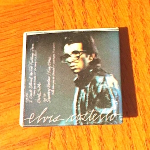 May include: A cassette tape case with a photo of a man wearing glasses and a black leather jacket. The text "Elvis Costello" is printed on the case.