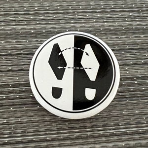 May include: A round, black and white button with a graphic of two footprints, one black and one white, facing each other. The footprints are separated by a dotted line with an arrow, suggesting a change of direction or a transition.