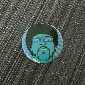 May include: A round button with a light blue background and a cartoon illustration of a man's face. The man has a mustache and goatee, and the text "BAD MOTHERFUCKER" is above his eyes.