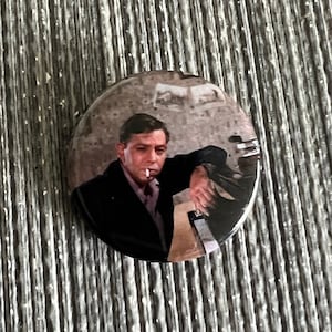 May include: A round button featuring a man in a black suit, smoking a cigarette. The image has a vintage aesthetic, with a blurred background. The button is set against a textured, gray background.