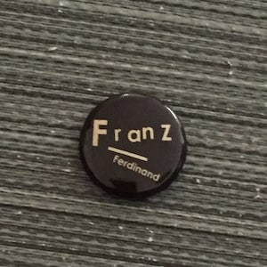 May include: A black round button with the text "Franz Ferdinand" in gold lettering.