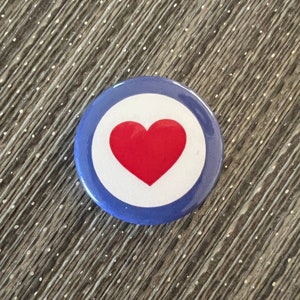 May include: A round button pin with a red heart in the center, surrounded by a white circle, and a blue outer ring. The pin is set against a textured, gray surface.