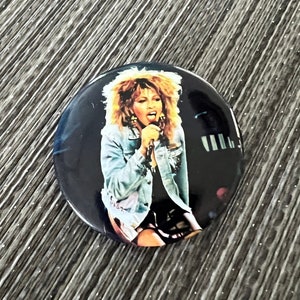 May include: A round button featuring a black and white photo of a woman with long, curly hair singing into a microphone. She is wearing a light blue denim jacket and a black skirt.