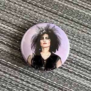 May include: A round button with a black and white image of a woman with dark hair and dark clothing. The background is a light purple color.
