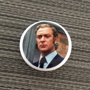 May include: A white button with a round image of a man with brown hair wearing a dark suit and a white shirt with a dark tie. The man is looking directly at the camera.