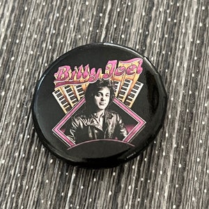 May include: A black button featuring a portrait of Billy Joel with the text "Billy Joel" in pink and yellow. The background includes a piano keyboard design and a pink diamond frame.