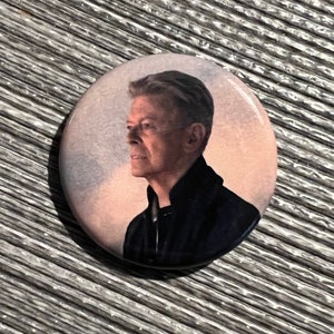 May include: A close-up of a round button featuring a black and white image of a man with short, gray hair. The man is looking to the left of the image. The background is a light pink and white.