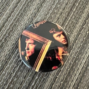 May include: A round button with a black and gold design featuring two men's faces. The men are looking in different directions and are framed by a gold frame.