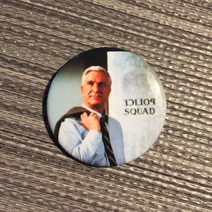 May include: A round button with a photo of a man wearing a blue shirt and a tie. The text "Police Squad" is printed on the button.