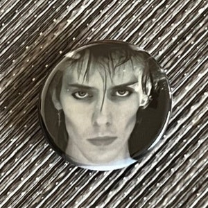 May include: A black and white button featuring a close-up portrait of a person with wet hair. The person is looking directly at the camera with a serious expression.
