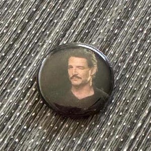 May include: A black and white button featuring a man with a beard and mustache wearing a black shirt.