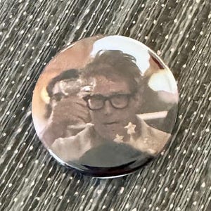 May include: A round button featuring a close-up image of a man wearing glasses and a shirt with stars. The man is holding a glass of liquid.