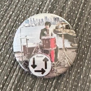 May include: Round button featuring a drummer playing a red drum set with a cityscape backdrop. The button has a black and white logo with arrows and the word "Premier". The button is on a gray and white dotted surface.