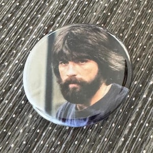 May include: A round button with a photo of a man with a beard and long hair. The man is wearing a blue shirt and has a serious expression on his face.