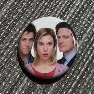 May include: A round button featuring a photo of three people. The people are a man with brown hair, a woman with blonde hair, and a man with dark hair. The woman is in the center of the button and is looking at the camera with a surprised expression. The men are on either side of the woman and are looking at the camera.