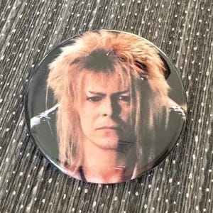 May include: A round button featuring a close-up portrait of David Bowie as Jareth the Goblin King from the movie Labyrinth. Bowie is wearing a black leather jacket and has long, blonde hair.