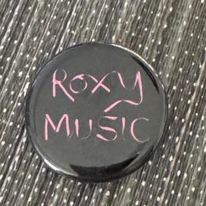 May include: A black button with the words "Roxy Music" in pink script. The button is round and has a glossy finish. The background is a gray and white dotted fabric.
