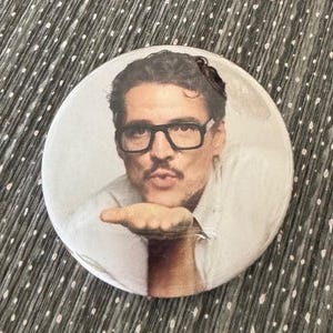 May include: A round button featuring a portrait of a man with glasses blowing a kiss. The image is printed on a white button with a glossy finish. The button is a fun accessory.